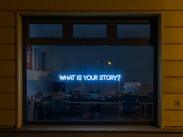 A neon window sign that says "what's your story?"